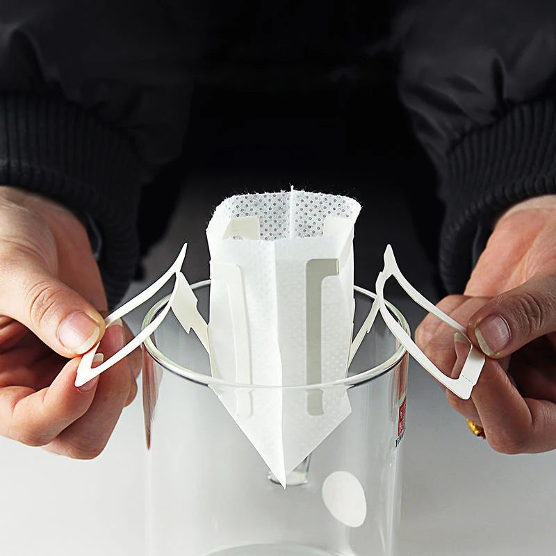 Hands opening a disposable drip coffee filter bag with hanging ear tabs
